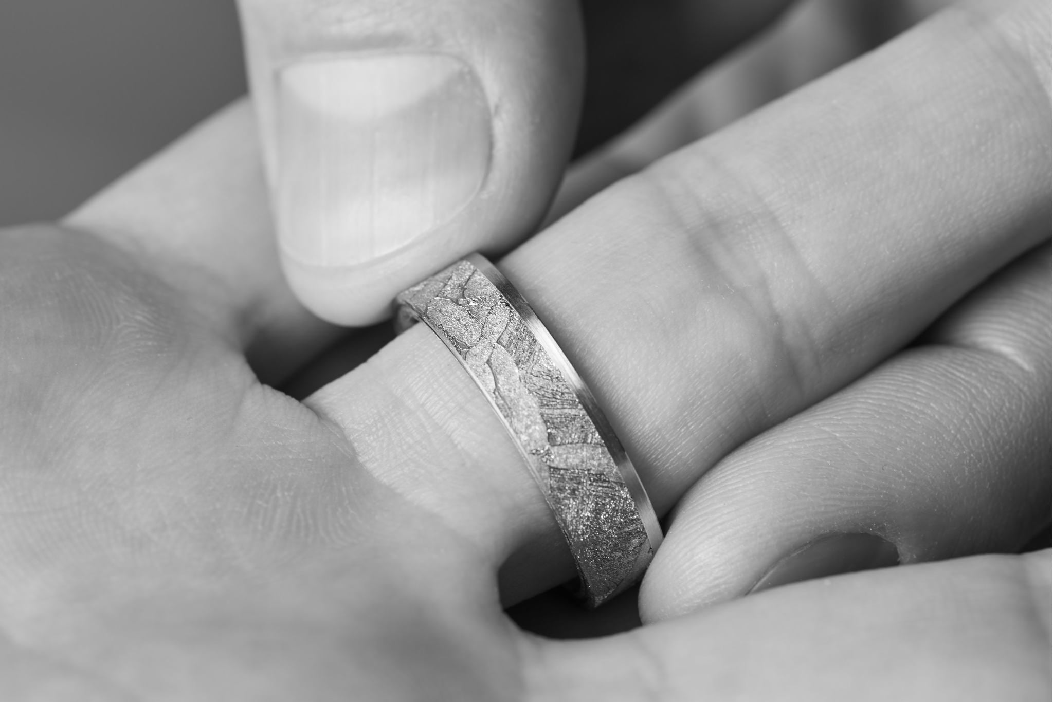  Breiter Ring aus Meteorit mit wunderschönem metallisch glänzenden Widmanstätten Muster. Innenseite aus Gold. Seitlich eckige Kanten. An einer Seite verläuft ein Band aus Gelbgold neben dem Meteoriten-Band. Scherzweise Foto, wo ein Mann einer Frau den Ring an die Hand steckt.