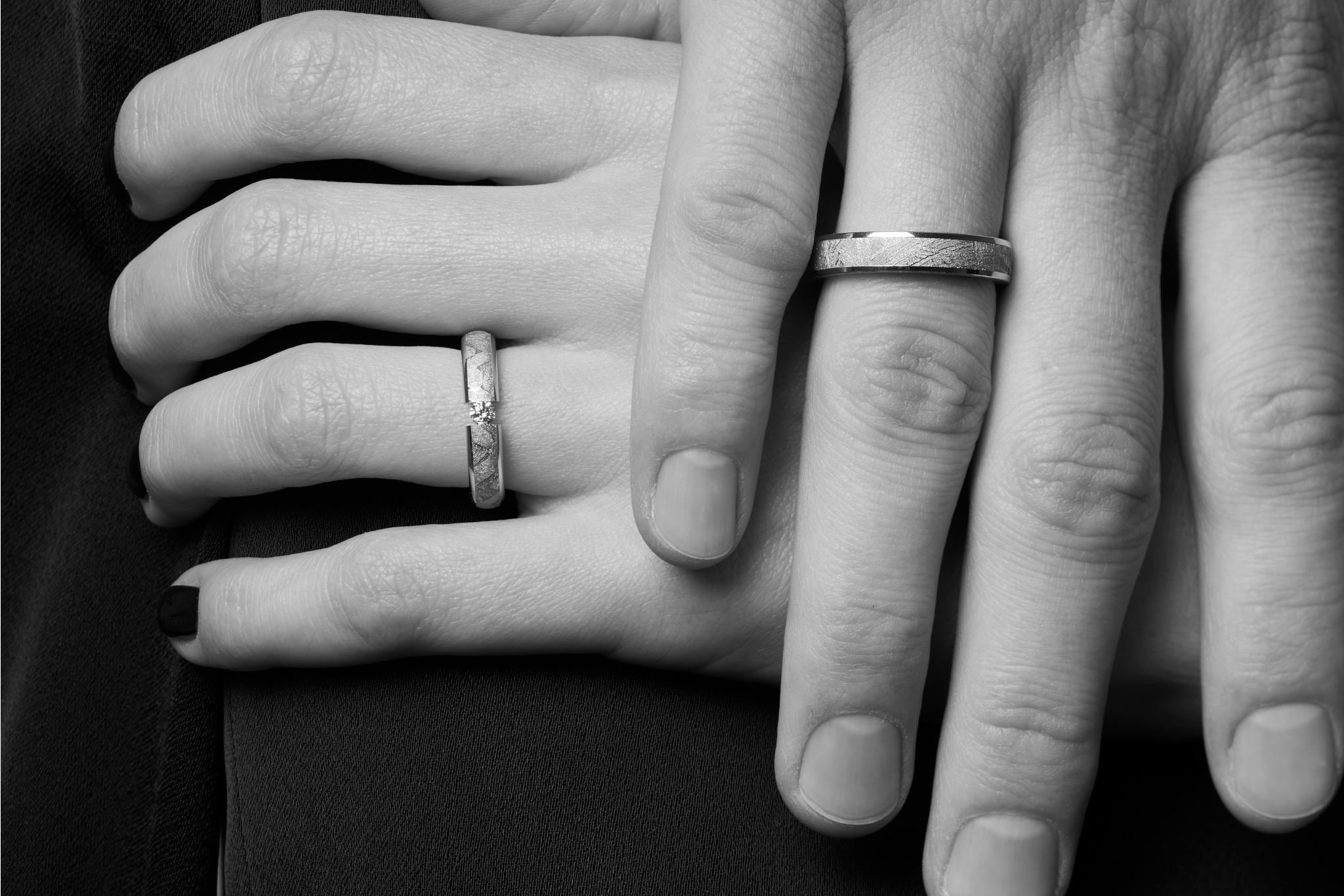 Ring aus Meteorit mit wunderschönem metallisch glänzenden Widmanstätten Muster. Innenseite aus Gold. Oben leicht abgerundet, seitlich jeweils ein Goldband das an der Oberseite poliert ist und seitlich matt gebürstet. Der Ring wird von einer Männerhand getragen, die auf einer Frauenhand liegt. Schwarz-weiss Foto