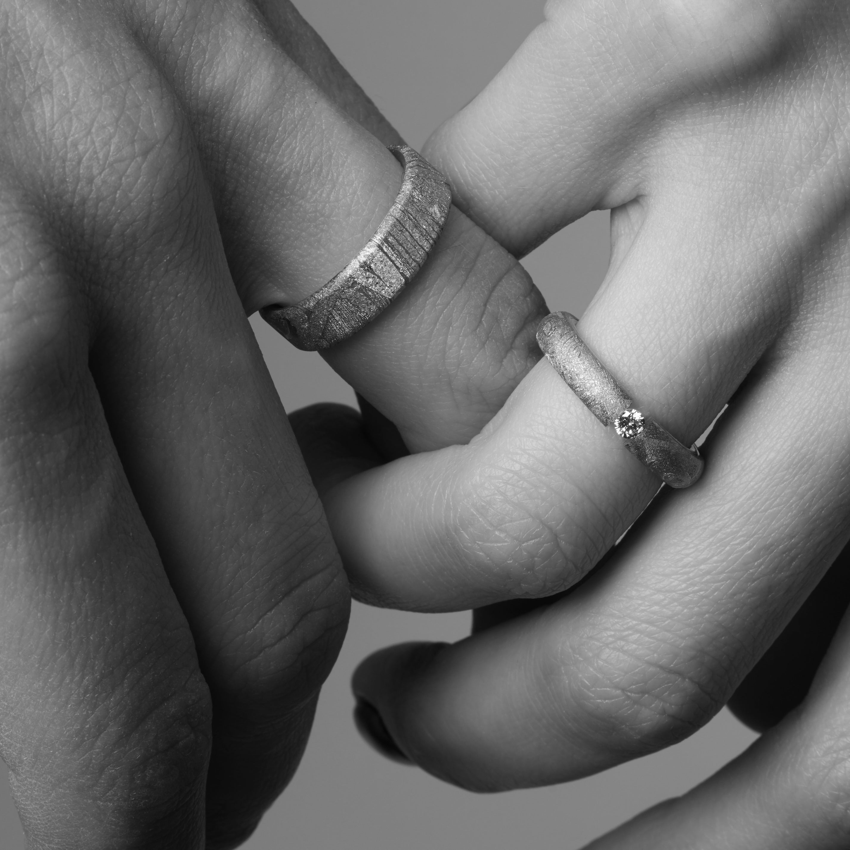 Die Hände eines Paars, das sich die Hand hält. Beide tragen einen Meteoriten Ring. Paarringe. Ihrer ist mit einem Brillant besetzt.